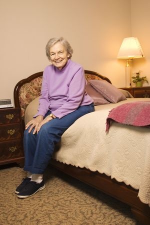 A senior woman wearing a purple sweater and blue plants smiles as she sits on her bed