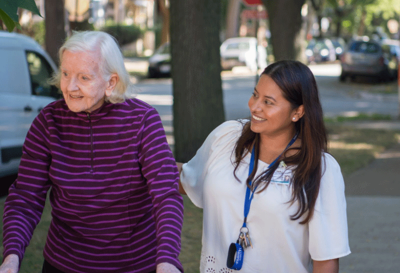 A senior female walks with the assistance of a walker. A young female caregiver accompanies her.