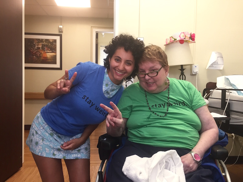 A young women and an older woman in a wheel chair. Both pose for a photo, throwing the "peace sign" with their right hands and both wearing a shirt with text across the chest that reads: "stay weird"