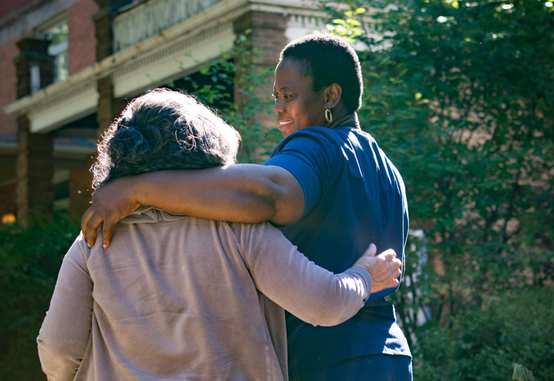 Two friends walk arm in arm on a neighborhood street.