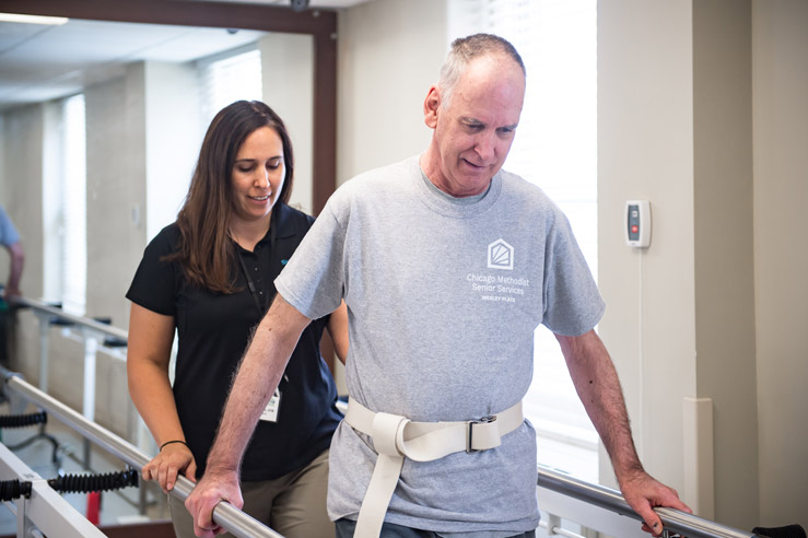 A man takes part in a walk exercise therapy exercise as a caregiver follows closely behind him