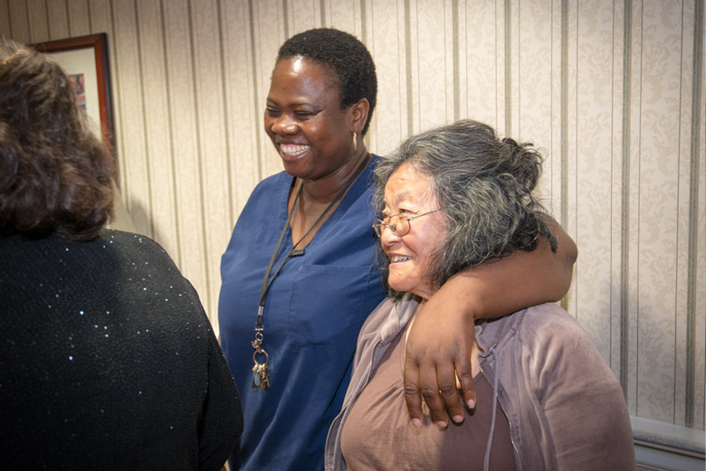 A caregiver embraces an older woman with an arm around her shoulder.