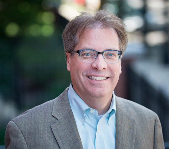 A man with short, light brown hair and glasses and wearing a blue shirt with a gray suit jacket, smiles.