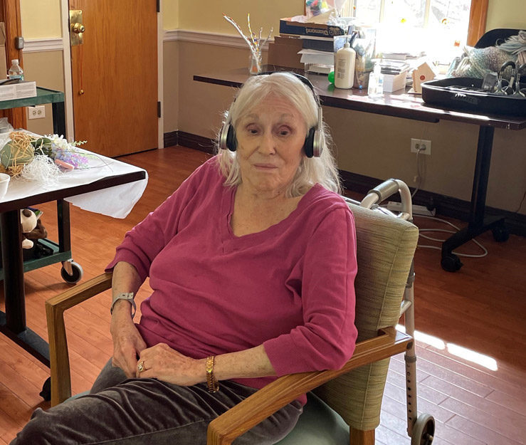 An elderly woman in a pink shirt and gray pants, listens to something through her headphones