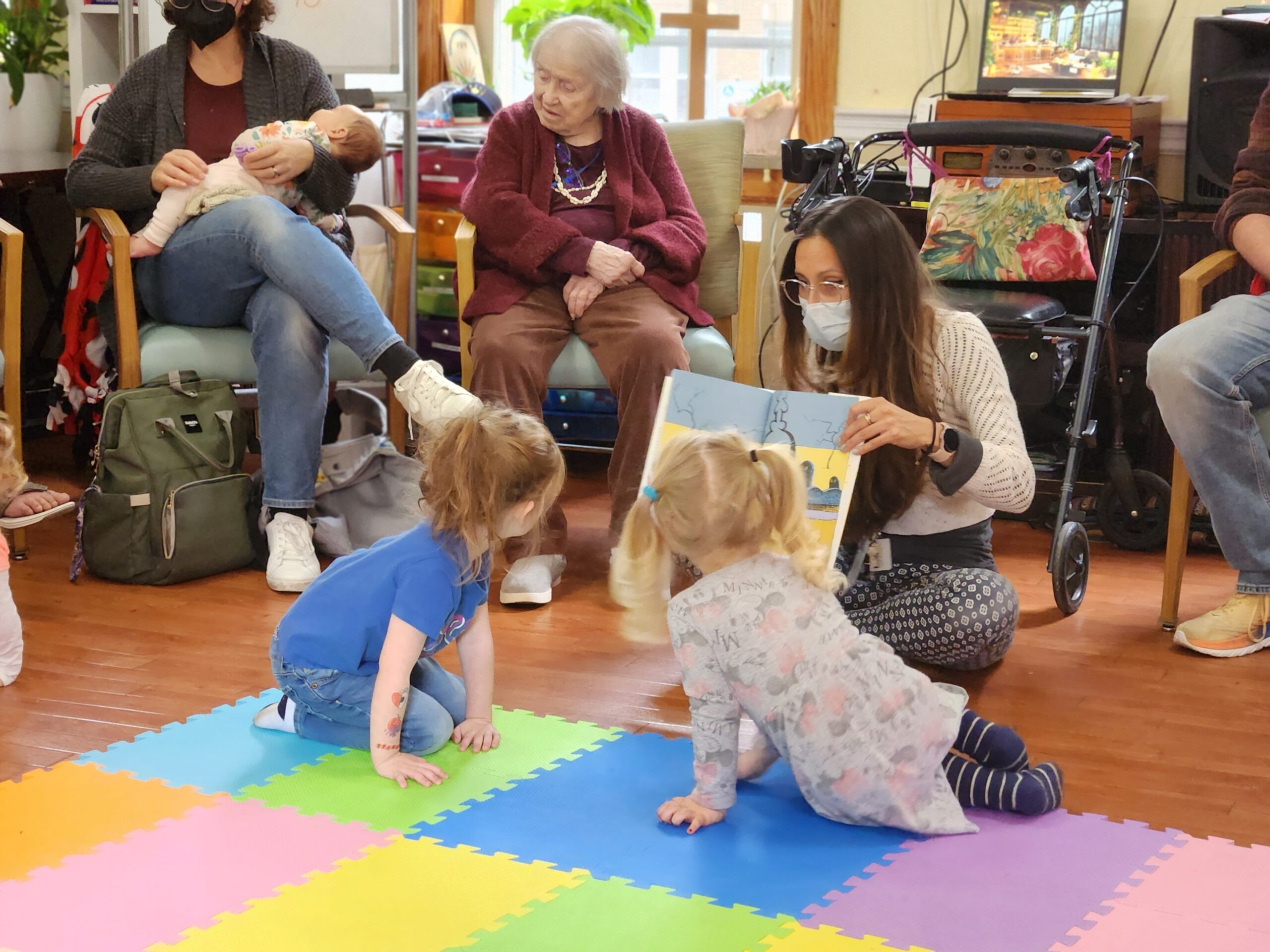 Two children sit on floor listening to story time. Adults and seniors are seated behind them.