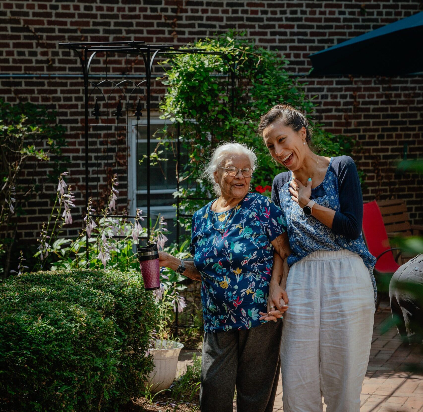 A young caregiver and older woman walk together, holding hands and laughing in a green garden.
