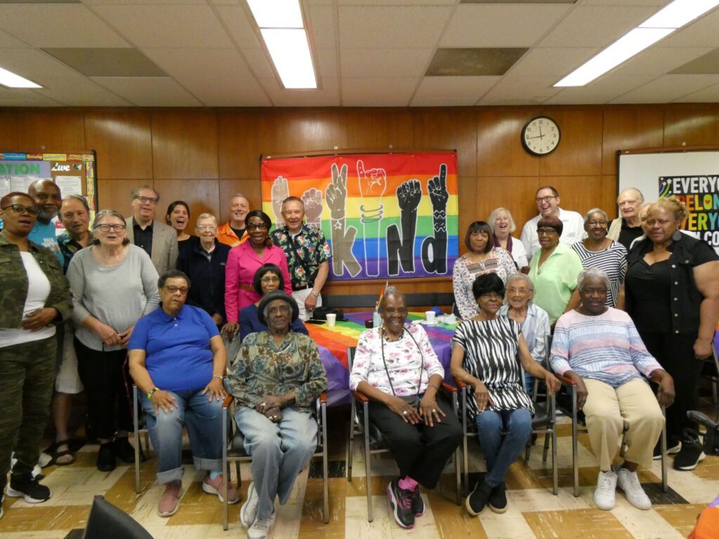 A large group of senior sit in front of a pride flag with the words "Be Kind" written on it.