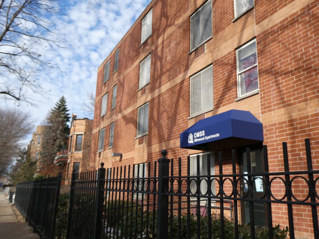 A red brick apartment building with a blue awning and a fence around the yard.