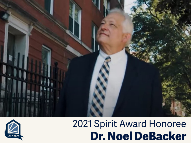 A man in a suit and tie walks down the sidewalk and gazes up at a brick building.