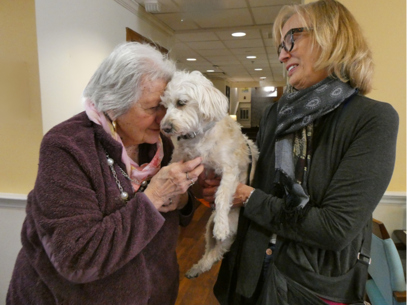 An woman holds a small, white dog, while an older woman pets the dog and puts her forehead against it.