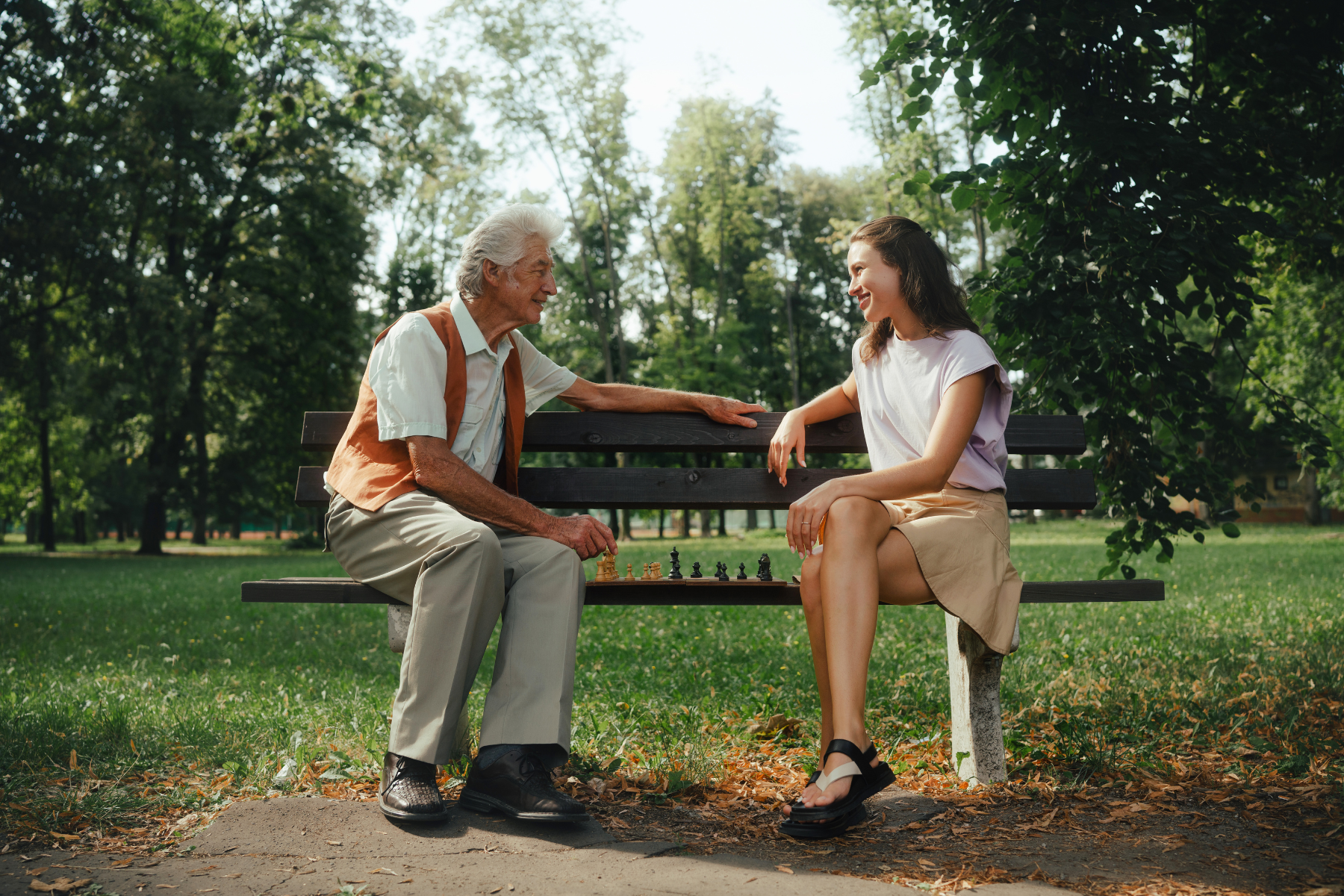 An older man and young woman talk together on a park bench in a green park.