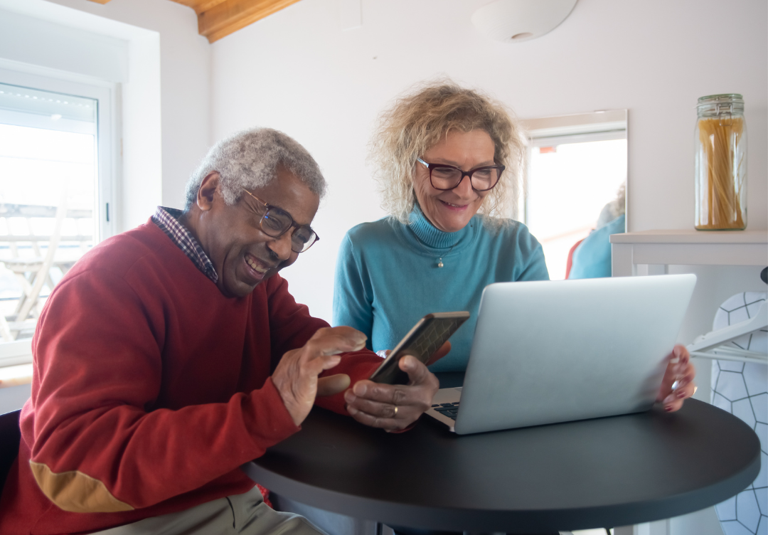 An senior man texts on his cell phone next to a senior woman typing on the computer.