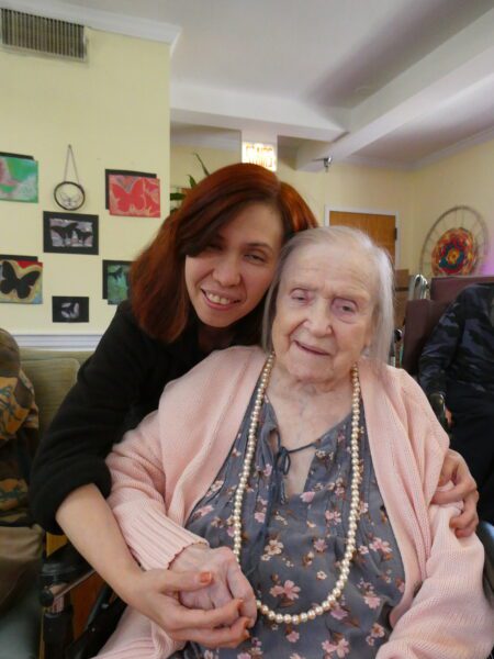 A nurse stands behind an older woman in a wheelchair and leans on her shoulder, they both smile for the camera.