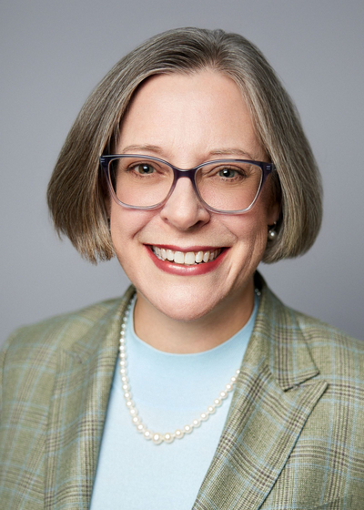 Portrait photo of a woman with short grey hair and glasses in a green blazer, smiling.