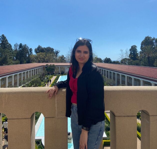 A young woman stands in front of a historic building and a bright blue sky.