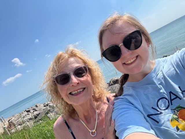 An older woman and a younger woman wearing sunglasses smile for a selfie by a lake.