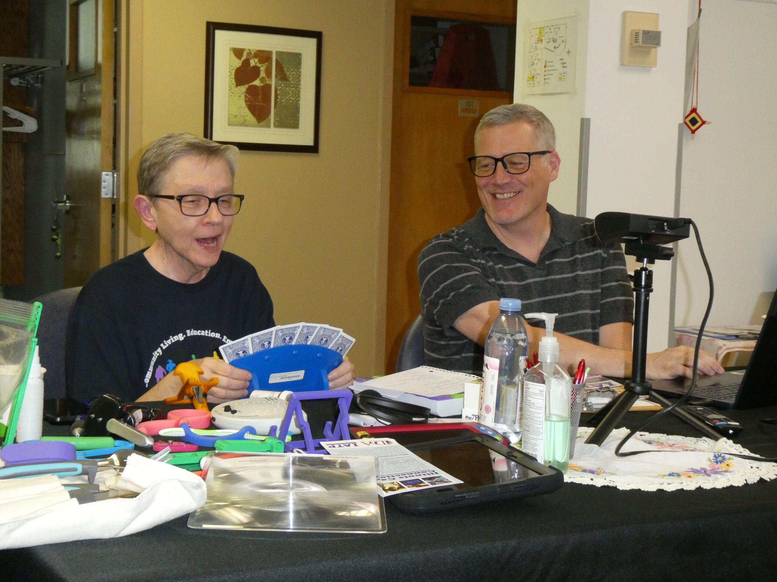 A presenter shows an assistive technology playing card holder, on a table with other devices.