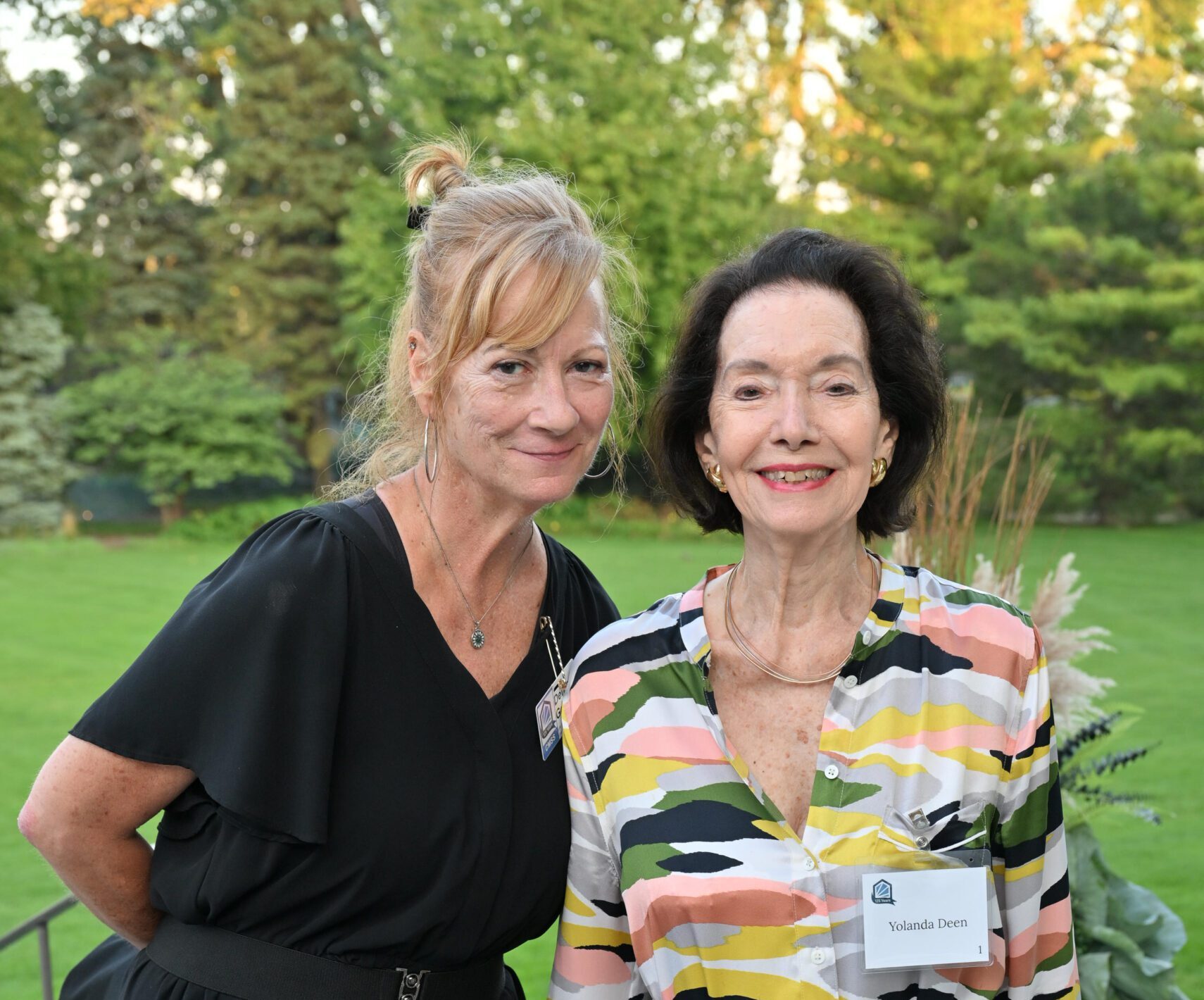 A woman with red hair and a woman with black hair pose in cocktail attire on a patio with a green yard behind them.