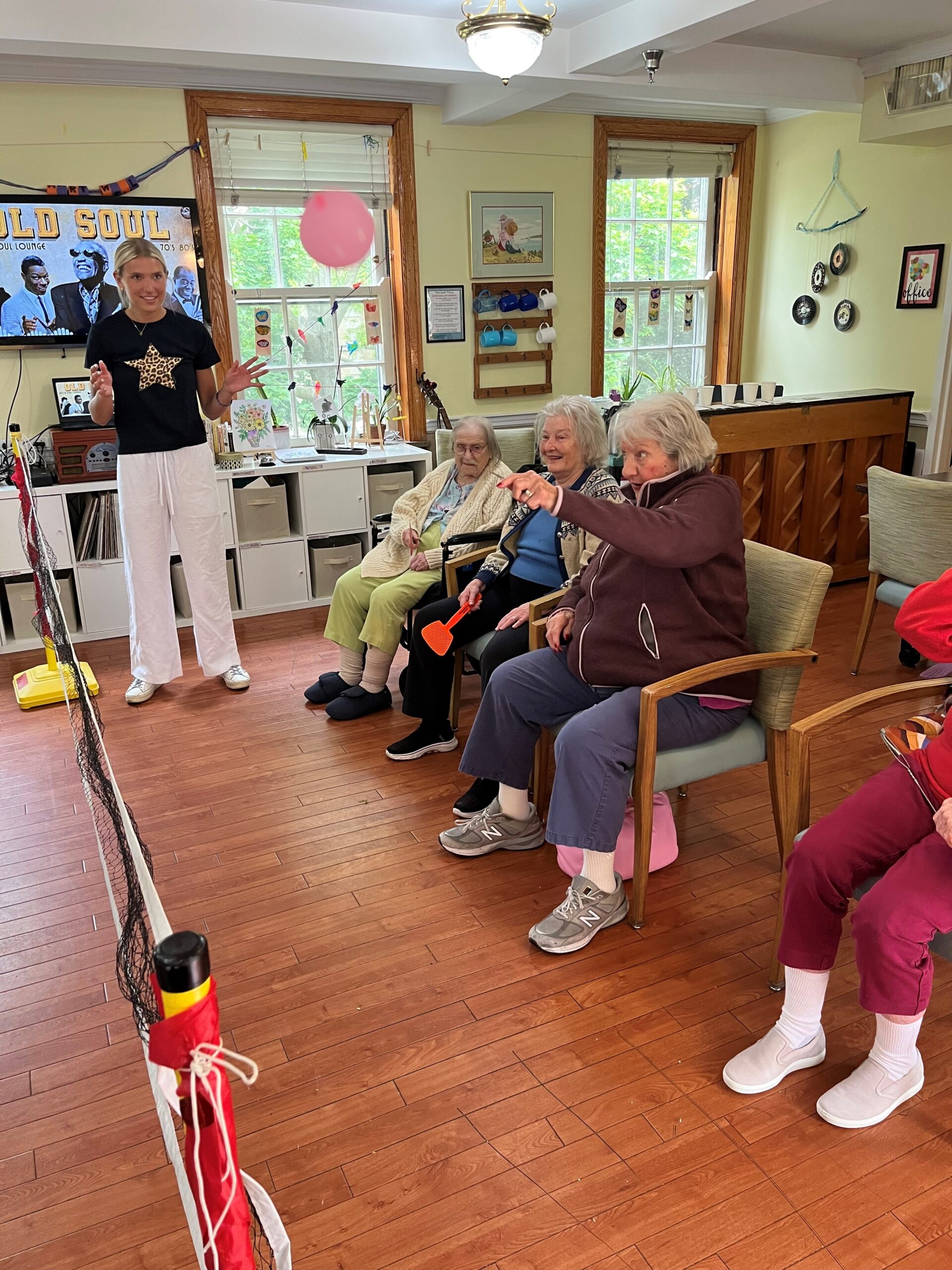 A young woman tosses a pink balloon in the air to volley with three older women seated near her. Everyone is smiling.