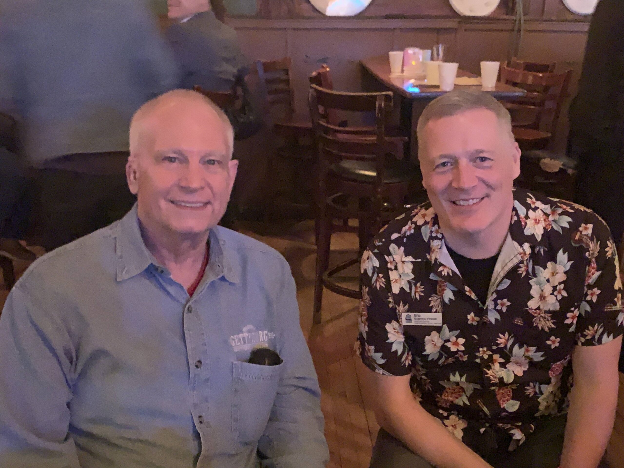 Two men in button-up shirts seated in a dim bar.