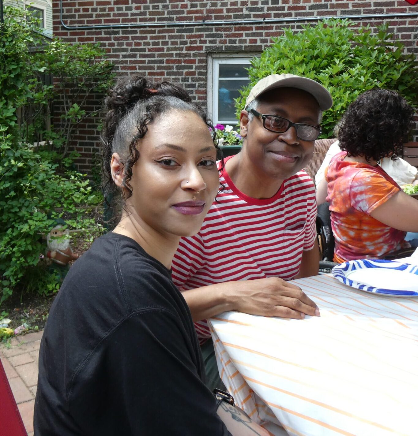 A woman with her curly hair in a bun and a man in a striped shirt and hat sit at a table with a red umbrella on a patio.