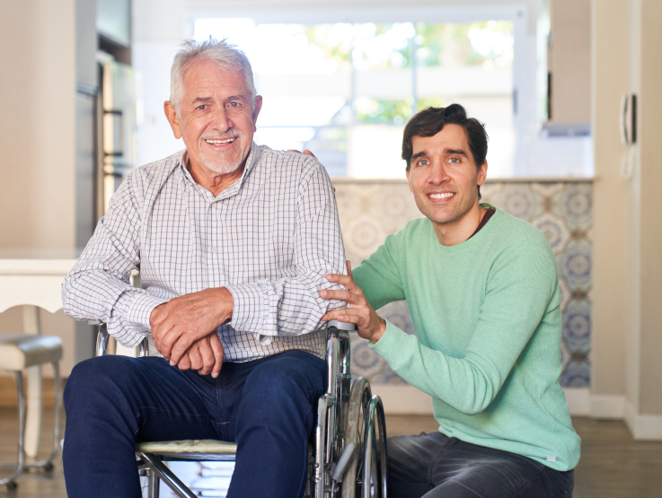A young man crouches down next to an older man in a wheelchair. The two are intergenerational friends, part of the Senior Connections program.